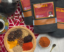 A bowl filled with spices, coffee beans, and peppers rests on a patterned mat, capturing the essence of rich Middle Eastern flavors. Nearby are packets of World Spice Kaffe Baharat—a unique coffee-infused blend from World Spice—accompanied by a steaming cup of coffee, a teapot, and a small bowl showcasing an orange spice blend.