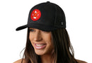 A woman with long brown hair smiles while wearing the Haka Hat Kansas City Trucker Hat in black, featuring a red and gold KC X patch. The black snapback by Haka Hat stands out against the white background.