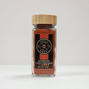 A glass jar with a bamboo lid, labeled "Spice Wheel Kashmiri Chili," sits on a white surface against a plain white background. The jar contains finely ground Kashmiri chili powder from Spice Wheel, celebrated for its vibrant red color and mild heat.
