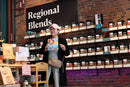 A person wearing a pink hat and black coat smiles while holding a World Spice cookbook in front of a display of spice jars labeled “Regional Blends”—featuring enticing options like World Spice Pink Chili Crisp and pink peppercorns—inside a store with brick walls.