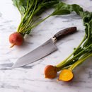 The Messermeister Kawashima 8" Chef's Knife rests on marble between a whole golden beet with greens and a sliced beet, showcasing its vibrant yellow interior and fresh tops.