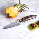 A halved pineapple with its top sits on marble next to a Messermeister Kawashima Paring Knife - 3.5". Kiwi slices and pomegranate seeds are nearby.