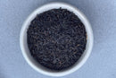 A white bowl filled with loose World Spice Keemun tea leaves from World Spice, renowned for their distinctive smoky black tea aroma, sits on a light gray surface.