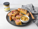 A plate of sliced sausage rolls topped with seeds, a bowl of dipping sauce, a knife, a gray-striped napkin, and a jar of Terrapin Ridge Farms Smokey Onion Gourmet Mustard by Terrapin Ridge Farms on a light surface.