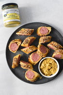 Sliced sausage rolls circle a bowl of gourmet mustard on a black plate, beside a jar of Terrapin Ridge Farms Smokey Onion Gourmet Mustard—the perfect condiment from Terrapin Ridge Farms—resting on a light gray surface.