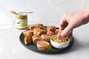 A plate of sliced sausage rolls with seasoning, one piece being dipped into a bowl of gourmet mustard. A jar of Terrapin Ridge Farms Smokey Onion Gourmet Mustard sits in the background on a light surface.