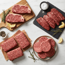 A selection from the Dude Food Kings Meat Box by Dude Food, featuring assorted raw American beef cuts such as strip steaks and ground beef patties, is arranged on plates and boards with herbs, lemon wedges, salt, and pepper atop a white marble surface.