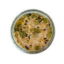 A glass jar of SALTOPIA Infused Sea Salts' Korean BBQ Salt, a sweet and savory blend with coarse salt, sesame seeds, black sesame seeds, dried green herbs, and yellow spices, viewed from above on a white background.