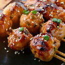 Close-up of glazed grilled chicken meatballs on skewers, topped with sesame seeds and green onions, finished with SALTOPIA Infused Sea Salts' Saltopia Korean BBQ Salt, served on a dark plate.