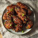 A plate of crispy Korean fried chicken wings glazed with sauce, garnished with sesame seeds, green onions, and a sprinkle of SALTOPIA Infused Sea Salts' Saltopia Korean BBQ Salt, sits on a cloth napkin.