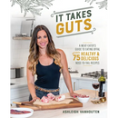 A smiling woman stands in a kitchen behind a wooden cutting board with cuts of meat, featured in It Takes Guts: A Meat-Eater’s Guide to Eating Offal with Over 75 Delicious Nose-To-Tail Recipes - Paperback by Ashleigh Vanhouten.