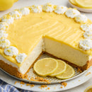 A creamy lemon cheesecake made with SALTOPIA Infused Sea Salts’ Saltopia Lemon Cheesecake Sugar: ZEST FOR LIFE, featuring a graham cracker crust, whipped cream, and lemon zest, with a slice missing and three lemon slices on the plate.