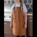 A person in a white chef's jacket and the Messermeister Vintage Amber Leather Apron stands in a modern kitchen, in front of a stainless steel grill and dark hexagonal-tiled backsplash.