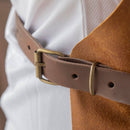 A close-up of the Messermeister Vintage Amber Leather Apron, featuring brown leather and a brass buckle, cinches a white textured shirt—showcasing Messermeister's timeless craftsmanship.