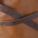 Close-up of brown leather straps, one embossed with Messermeister, resting on the textured surface of the handmade Messermeister Vintage Amber Leather Apron.