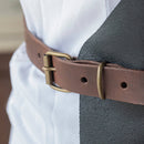 A close-up of the Messermeister Vintage Black Leather Apron, featuring a brass buckle and vintage design, worn over a white textured shirt and dark vest.