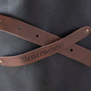 Close-up of two brown straps from the Messermeister Vintage Black Leather Apron crossed over a black surface, with "Messermeister" on one strap and brass rivets securing the ends, highlighting classic craftsmanship.