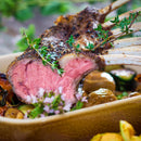 A close-up of medium-rare rack of lamb, garnished with fresh herbs and SALTOPIA Infused Sea Salts' Saltopia Herbs De Provence Seasoning Salt, served with roasted vegetables. The juicy meat has a pink center, browned crust, and a sprinkle of sea salt.