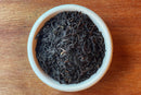 A white bowl filled with loose World Spice Lapsang Souchong tea leaves, a distinctive pine-smoked tea from Fujian, China by World Spice, sits on a wooden surface.