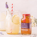 A jar of Honey Truck Co Lavender Infused Honey rests beside a mason jar of iced lemonade with lemon slices and a red striped straw. Lavender and flowers are visible in the background.