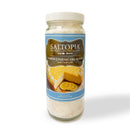 A clear glass jar with a white lid holds SALTOPIA Infused Sea Salts' Lemon Cheesecake Sugar: ZEST FOR LIFE, featuring a blue label with lemon cheesecake art and the text “LEMON CHEESECAKE SUGAR - Zest for Life.”.