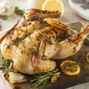 A whole roast chicken, seasoned with SALTOPIA Infused Sea Salts' Lemon Pepper Salt: PUCKER UP BUSTER and garnished with lemon slices, rosemary, and garlic, is served on a wooden cutting board with plates and utensils in the background.