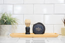 A shaving brush and a round Shave Soap Puck by The Immaculate Beard rest on a wooden tray, set against a backdrop of potted plants and white subway tiles—an ideal setup for anyone looking for gentle care for sensitive skin.
