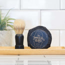 A shaving brush and a round bar of The Immaculate Beard shave soap sit on a wooden tray, complemented by a plant and white tile backsplash in the background—a perfect addition to any men’s grooming routine, especially when paired with Immaculate Natural Beard Balm For Men With Lightweight Hold from The Immaculate Beard for optimal beard conditioning.