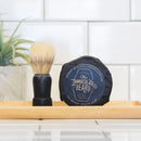 A shaving brush and a round bar of The Immaculate Beard Dark Shave Soap Puck rest on a wooden tray in front of a white tiled wall, with a green plant visible in the background.