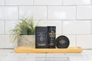 Three men’s beard care products from The Immaculate Beard, including the Conditioning Natural Beard Oil, are displayed in sleek black containers on a wooden tray, with a potted plant and white tiled wall in the background.