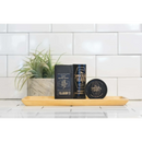 Three grooming products from The Immaculate Beard, including the Immaculate Fast Absorbing Beard Oil For Men Softens Skin, rest on a wooden tray in front of a potted plant, set against a white tiled background.