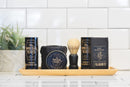 Grooming products, including natural beard care essentials like The Immaculate Beard’s Natural Beard Wash Bar, beard oil, beard balm, and a shaving brush, are neatly arranged on a wooden tray in front of white subway tiles and a small green plant.
