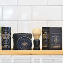 A wooden tray displays The Immaculate Beard’s beard oil, beard balm, beard wash, shaving brush, and an Immaculate Beard Dark Shave Soap Protective Lather for sensitive men in front of a white tiled wall.