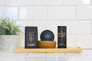 A wooden tray holds a potted plant, a box of The Immaculate Beard Natural Beard Wash Bar, a beard balm tin, a wooden bowl, and a bright beard oil bottle—offering the essentials for natural beard care—set against a white tiled wall.