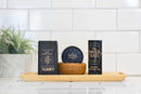 A wooden tray holds a potted plant, a bar of The Immaculate Beard Natural Wash Bar For Men Deep Clean, a tin of beard balm in a wooden bowl, and a bottle of bright beard oil, all set against a white tiled background.