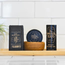 A bottle of Conditioning Natural Beard Oil from The Immaculate Beard, along with Dark Beard Wash and a tin of natural beard balm, are arranged on a wooden tray, with a small plant softly blurred in the background.