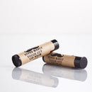 Two black lip balm tubes from The Immaculate Beard, with brown labels that read “Super Manly Tough Guy Lip Balm” and a mustache graphic. Perfect for including in the Shave Gift Set | The Immaculate Beard or pairing with natural shave soap for a classic hot shave, one tube stands upright while the other lies down, both reflected on a white surface.