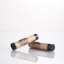 Two black tubes of The Immaculate Beard’s Super Manly Tough Guy Lip Balm, featuring brown labels with a mustache graphic and bold lettering, rest on a glossy white surface—one upright, one lying down. Crafted with natural ingredients and refreshing peppermint flavor, this lip balm is made especially for men seeking rugged style and serious protection.