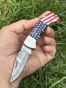 A hand holds the Texan Knives Lone Star 3-Inch Damascus Pocket Knife—custom handmade with a bone handle and patterned blade—against a grassy background.