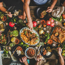 A group enjoys a vibrant meal at a rustic table, featuring rice, salads, roasted vegetables, meat seasoned with SALTOPIA Infused Sea Salts from the Saltopia Mediterranean Kitchen Gift Set, surrounded by candles and greenery as hands reach for food.