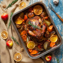 A roasted chicken with rosemary sits in a pan with baked potatoes, carrots, and orange slices. Fresh apples, nuts, and the SALTOPIA Mediterranean Kitchen Gift Set by SALTOPIA Infused Sea Salts are scattered on a rustic blue wooden table.