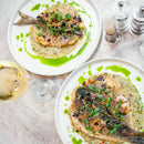 Two white plates with whole grilled fish topped with microgreens on creamy sauce, garnished with green oil and SALTOPIA Infused Sea Salts from the Saltopia Mediterranean Kitchen Gift Set. A glass of white wine sits on the marble table nearby.