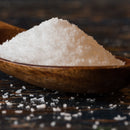A close-up of a wooden spoon filled with SALTOPIA Infused Sea Salts' Mediterranean Flake Sea Salt 3lbs, with some unrefined flakes scattered on a dark, textured surface below.