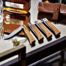 Four Messermeister Folding Steak Knives with wooden handles are lined up on a glass table beside a whiskey bottle, leather roll case, and tumbler. The first knife from the Messermeister set is open, showing its gleaming blade.