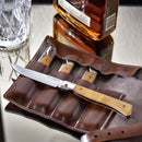 A set of four Messermeister Folding Steak Knives (4") with wooden handles rests in a brown leather case on a reflective table, next to a glass and a bottle of amber-colored liquor.
