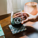 A hand holds a custom engraved Stemless Wine Glasses - Set of 4: The Classic with the letter K in a decorative border, resting on a patterned coaster.