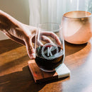 A hand holding a Swanky Badger Stemless Wine Glasses - Set of 4: Script, engraved with the letter K inside a scalloped border and resting on a square coaster.
