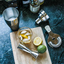 A Whiskey Essentials Bundle featuring a cocktail shaker, jigger, Hawthorne strainer, and a glass of whiskey with ice and a lemon wedge on a cutting board, displayed alongside a halved lemon and lime on leather coasters.
