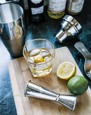 A Whiskey Essentials Bundle glass of whiskey on a leather coaster with ice and a lemon slice on a wooden board, surrounded by a cocktail shaker, a jigger, a lemon half, and a lime.