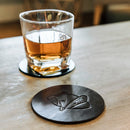 A glass of amber liquid sits on a round black leather coaster next to a matching coaster embossed with a badger logo, completing a Swanky Badger Whiskey Essentials Bundle.